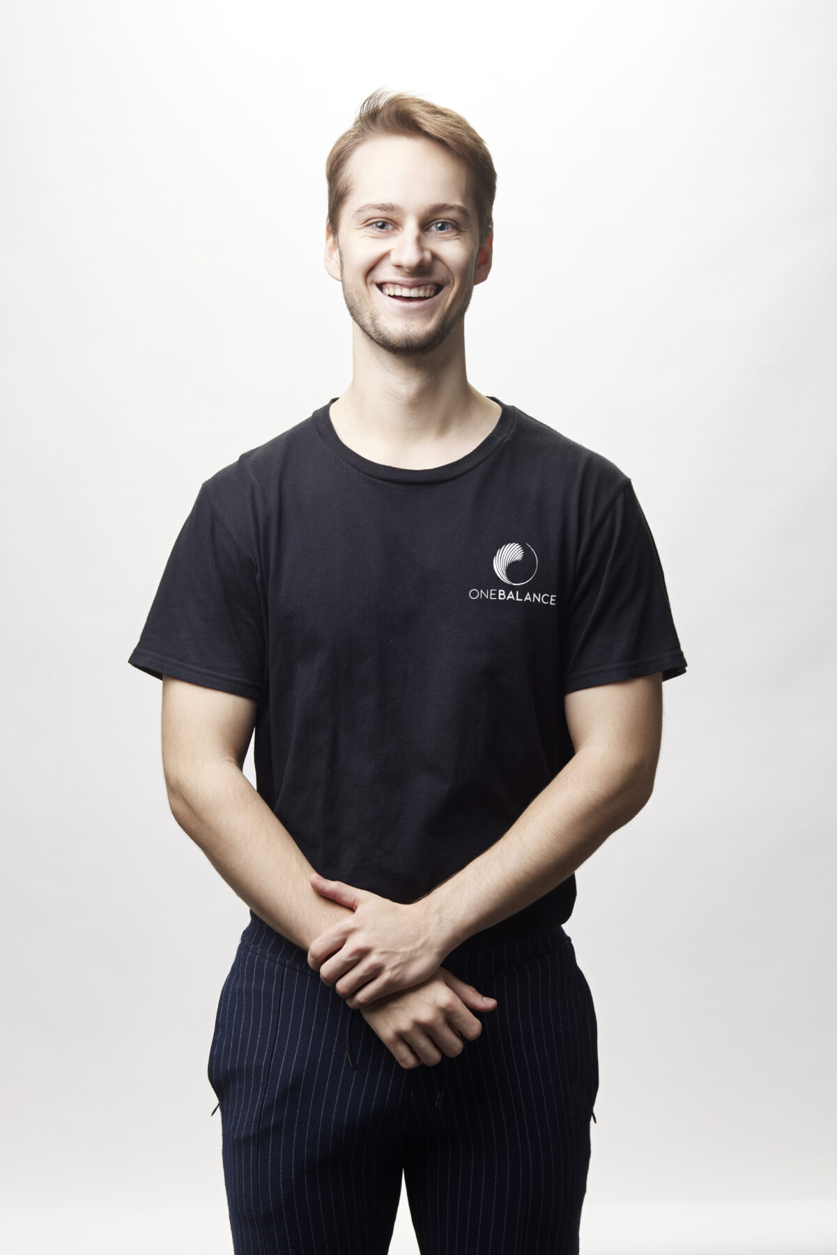 Lächelnder Mann im schwarzen T-Shirt steht vor weißem Hintergrund und hält die Hände locker vor dem Körper.